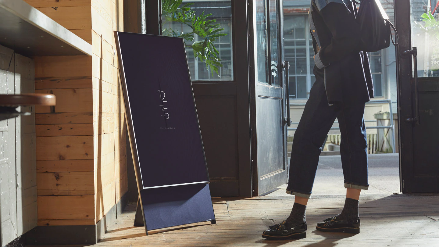 Un TV The Sero bleu marine est en Mode Portrait à l'entrée d'un café, montrant la météo au lieu d'avoir un écran noir. Un client est placé à côté.
